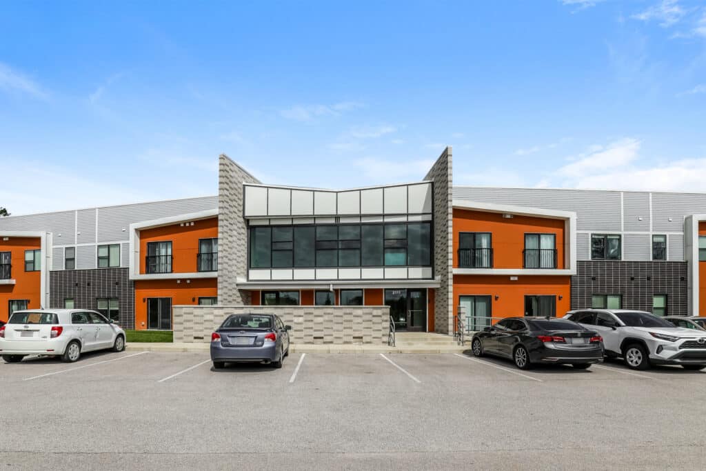 Modern two-story building with large glass entrance, orange and gray exterior accents, and cars parked in front in a paved parking lot under a blue sky.