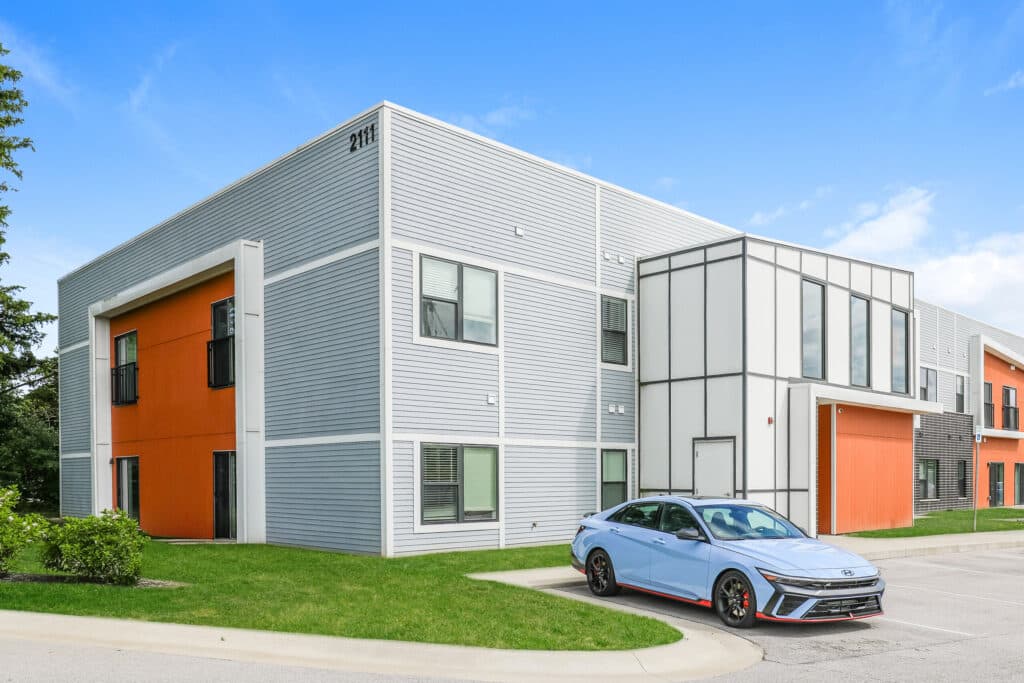 A modern two-story apartment building with gray and orange accents, large windows, and a parked light blue car in front under a clear blue sky.