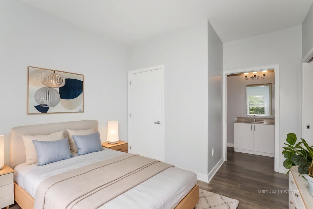 Modern bedroom with neutral tones, a bed with beige linens, nightstands with lamps, abstract wall art, and an adjoining bathroom visible through an open doorway.