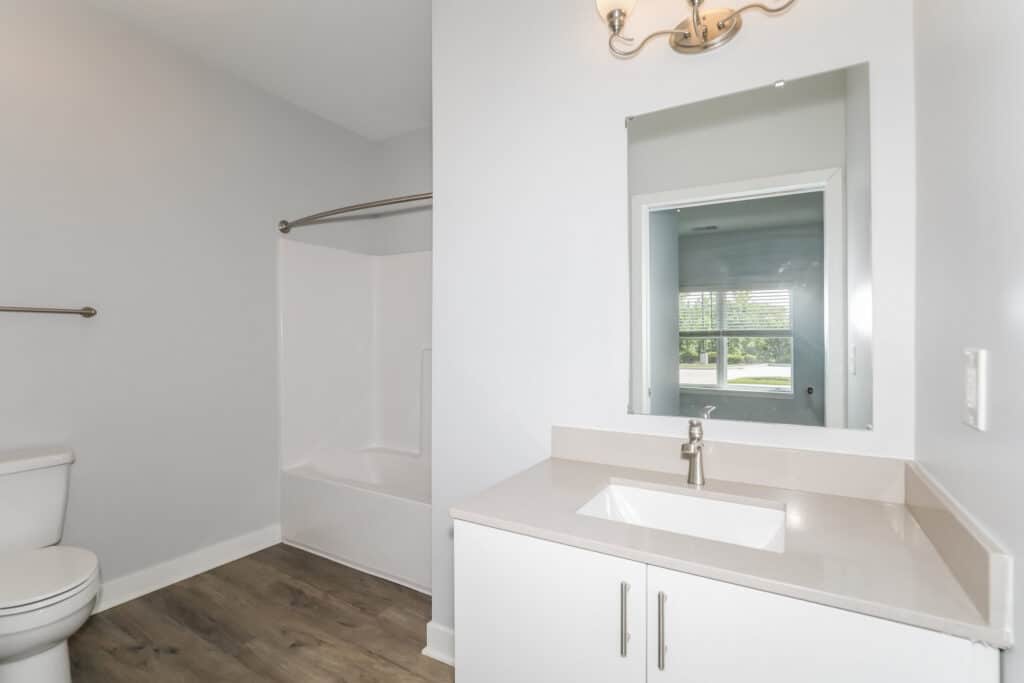 Modern bathroom with a white toilet, bathtub with shower, white vanity with sink, wall mirror, and overhead light fixture on light-colored walls and wood-style flooring.