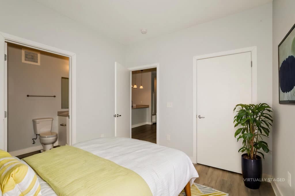 Bedroom with white walls, a bed with green and yellow bedding, a potted plant, two closed doors, and an open door showing a bathroom with a toilet. "Virtually staged" text in corner.