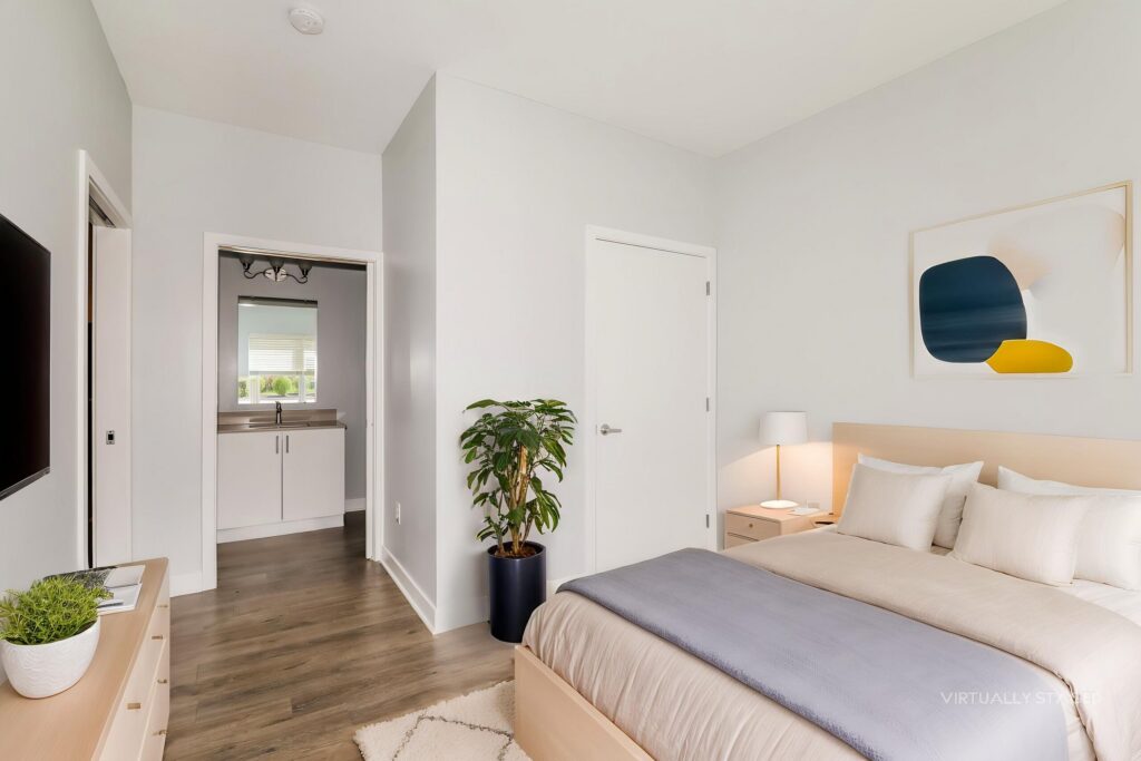 Modern bedroom with light wood furniture, a neatly made bed, a potted plant, and a view into an adjoining bathroom. Walls are white with minimal decor.