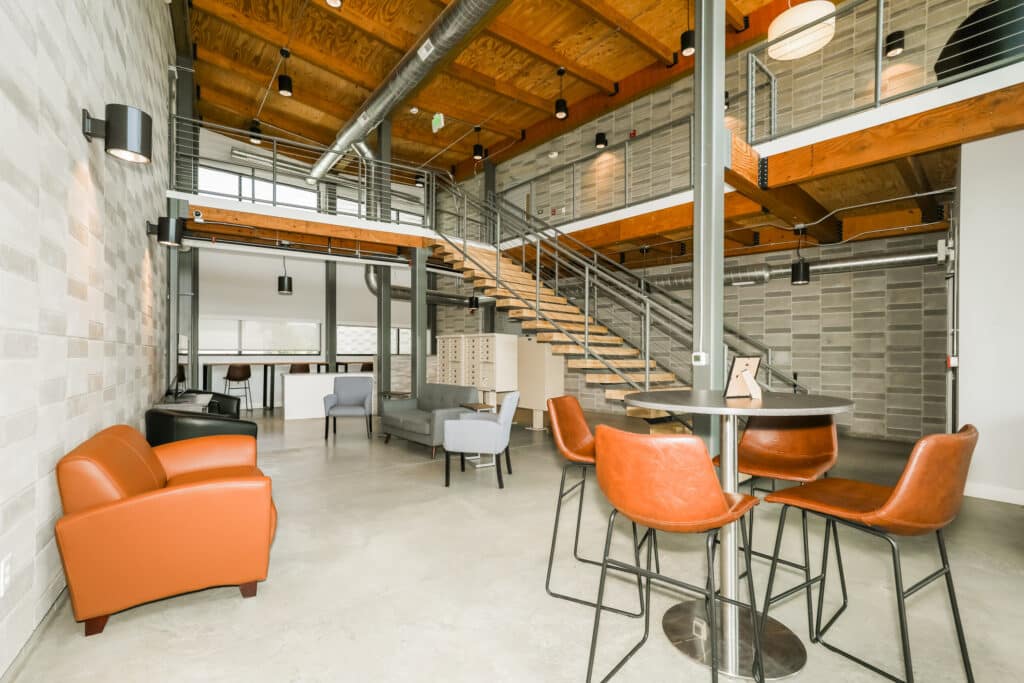 Modern open office lounge with leather chairs, bar stools, a round table, gray armchairs, exposed beams, metal staircase, and large windows.