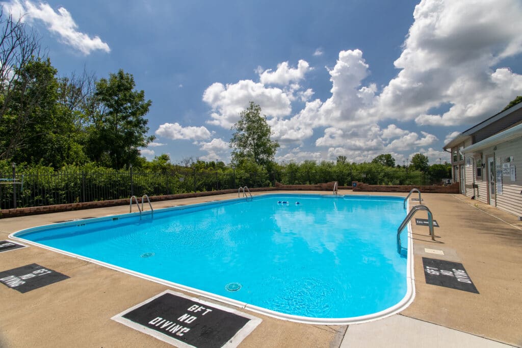 Rectangular outdoor swimming pool with clear blue water, surrounded by a concrete deck, metal ladders, safety signage, and greenery under a partly cloudy sky.
