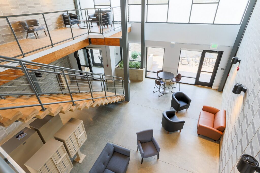 Modern two-story office lobby with seating areas, large windows, stairs, mailboxes, and natural light filling the open space.