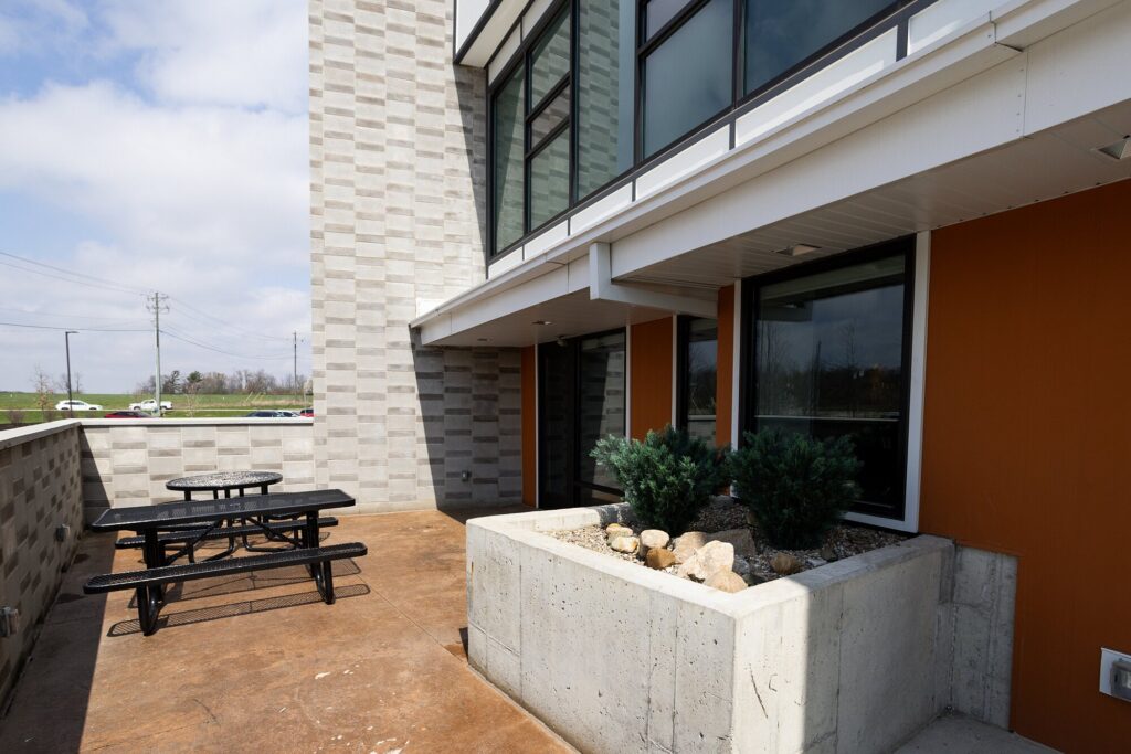 Outdoor seating area with black picnic tables, concrete planter with rocks and shrubs, and modern building exterior.