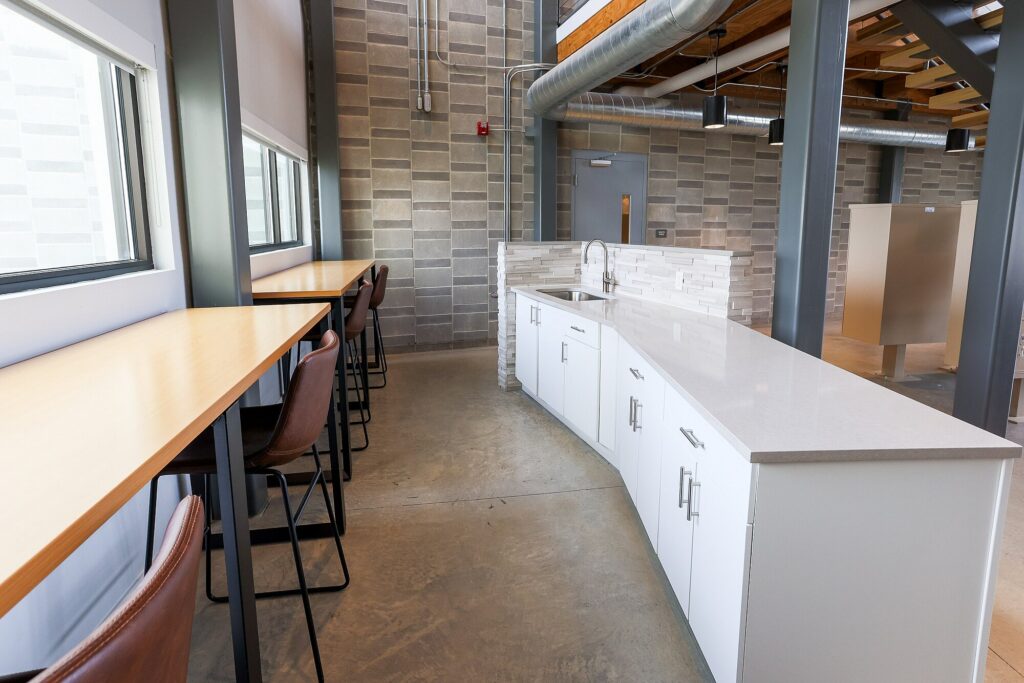Modern office kitchen area with white cabinets, a long counter, barstools, large windows, exposed ducts, and industrial-style brick walls.