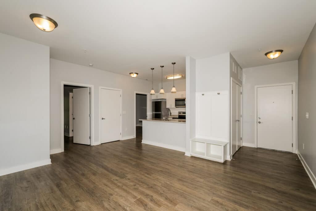 Unfurnished apartment interior with wood flooring, white walls, open kitchen with pendant lights, multiple doors, and a built-in entryway bench.