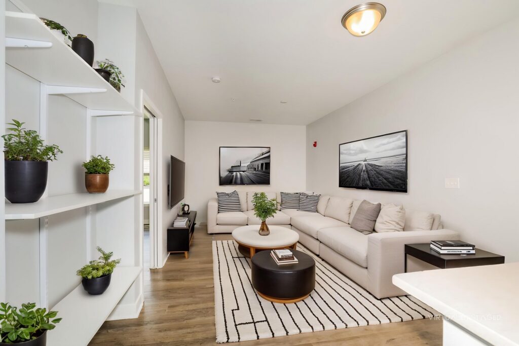 Modern living room with a beige sectional sofa, striped rug, coffee table, wall art, and shelves with potted plants. Light walls and wood flooring create a bright, clean atmosphere.